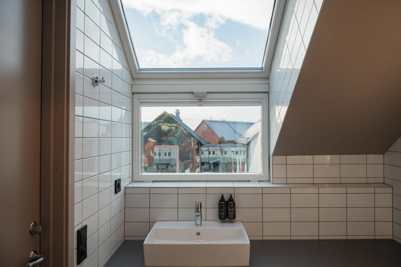 Et lyst, flisebelagt badeværelse med en vask, et vindue med udsigt over bygninger og et ovenlysvindue, der afslører himlen ovenover.