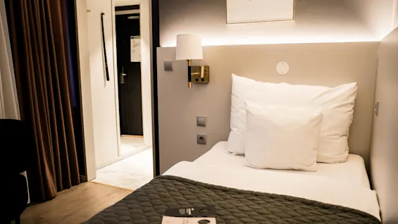 En prydligt bäddad säng med vita kuddar finns i ett hotellrum. Ett vadderat täcke täcker den. En broschyr ligger på sängen. Gardiner täcker ett fönster i närheten.
