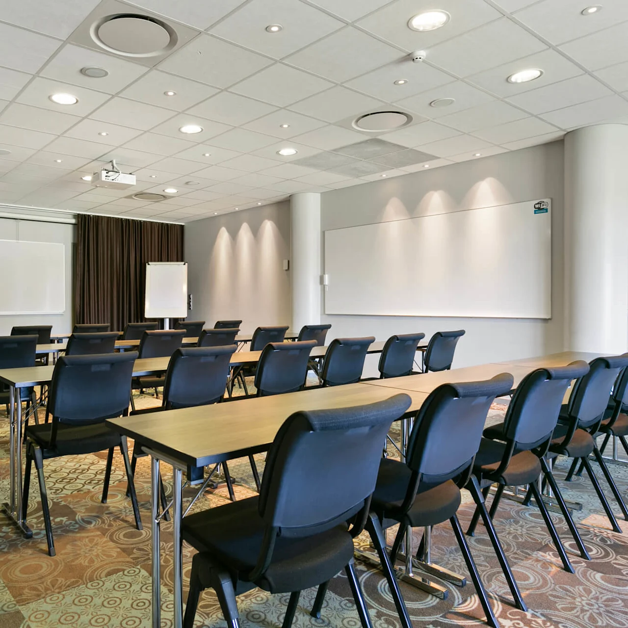 Klasselokale med rækker af sorte stole og borde, der vender mod et whiteboard, projektor hænger fra loftet; mønstret tæppe og vægge med gardiner skaber et formelt læringsmiljø.
