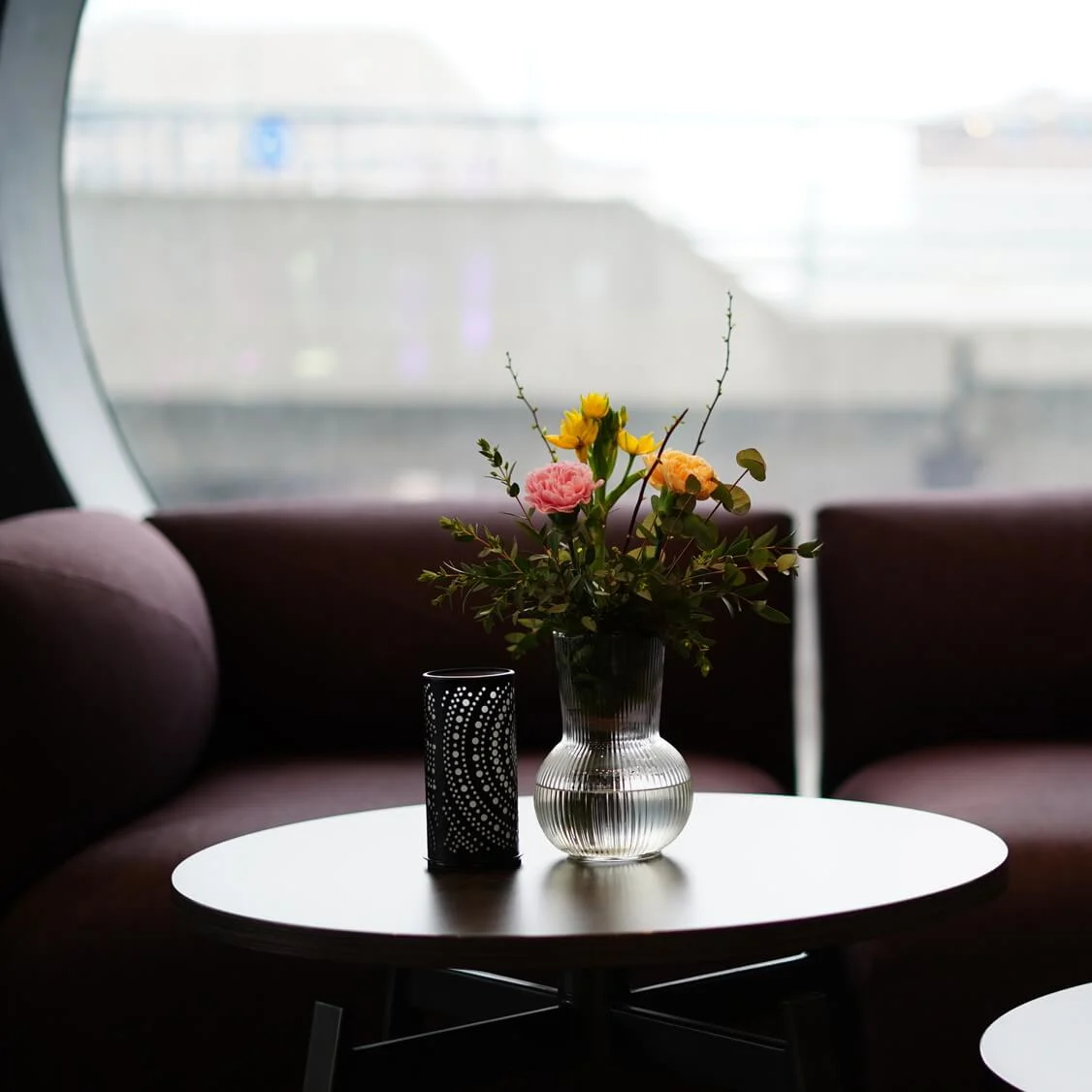 En vase med farverige blomster står på et rundt bord mellem to brune sofaer, i et rum med et stort rundt vindue og lilla lys.