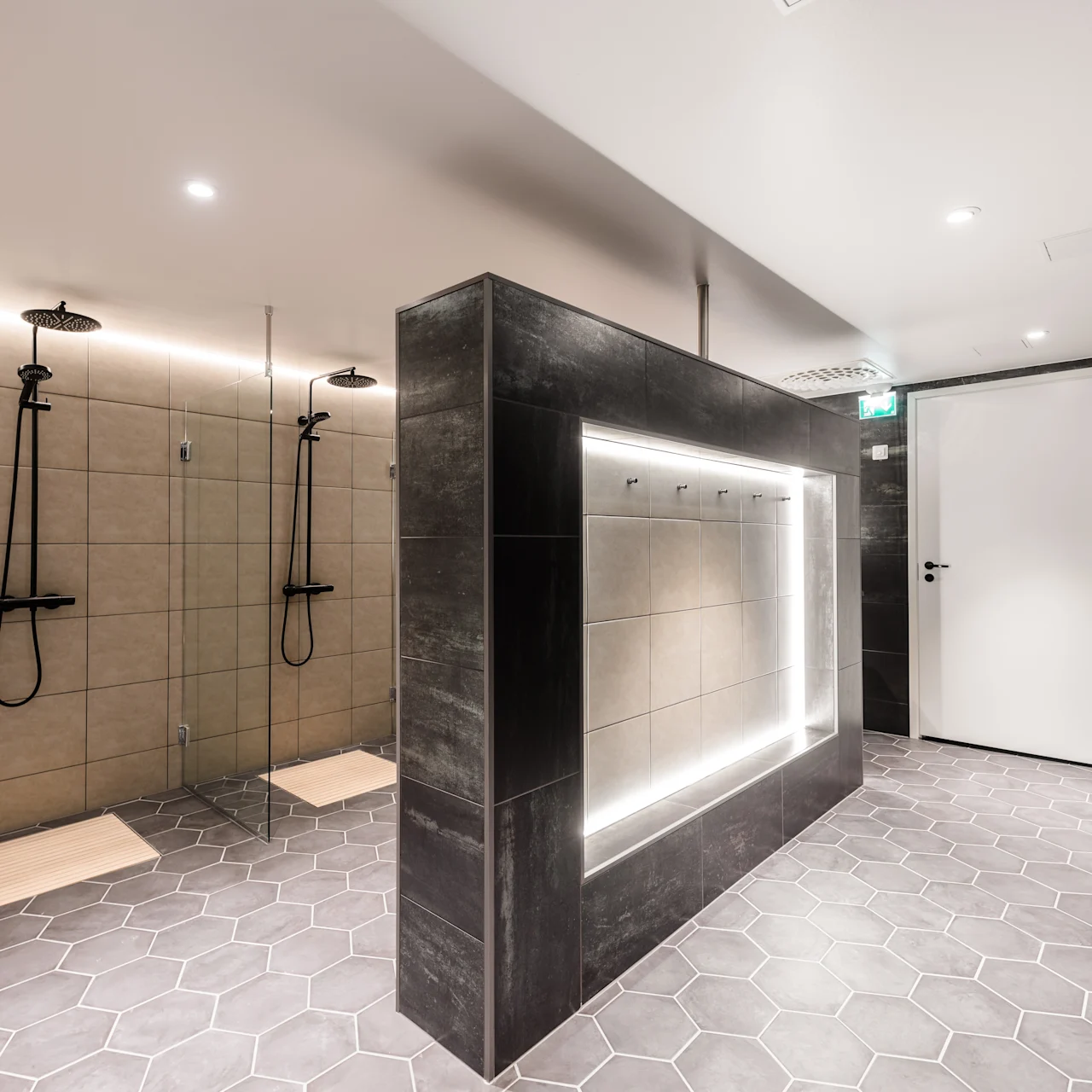 A modern shower area features two adjacent rain shower heads on tiled walls, with wooden mats below. The space includes hexagonal floor tiles and dim overhead lighting.