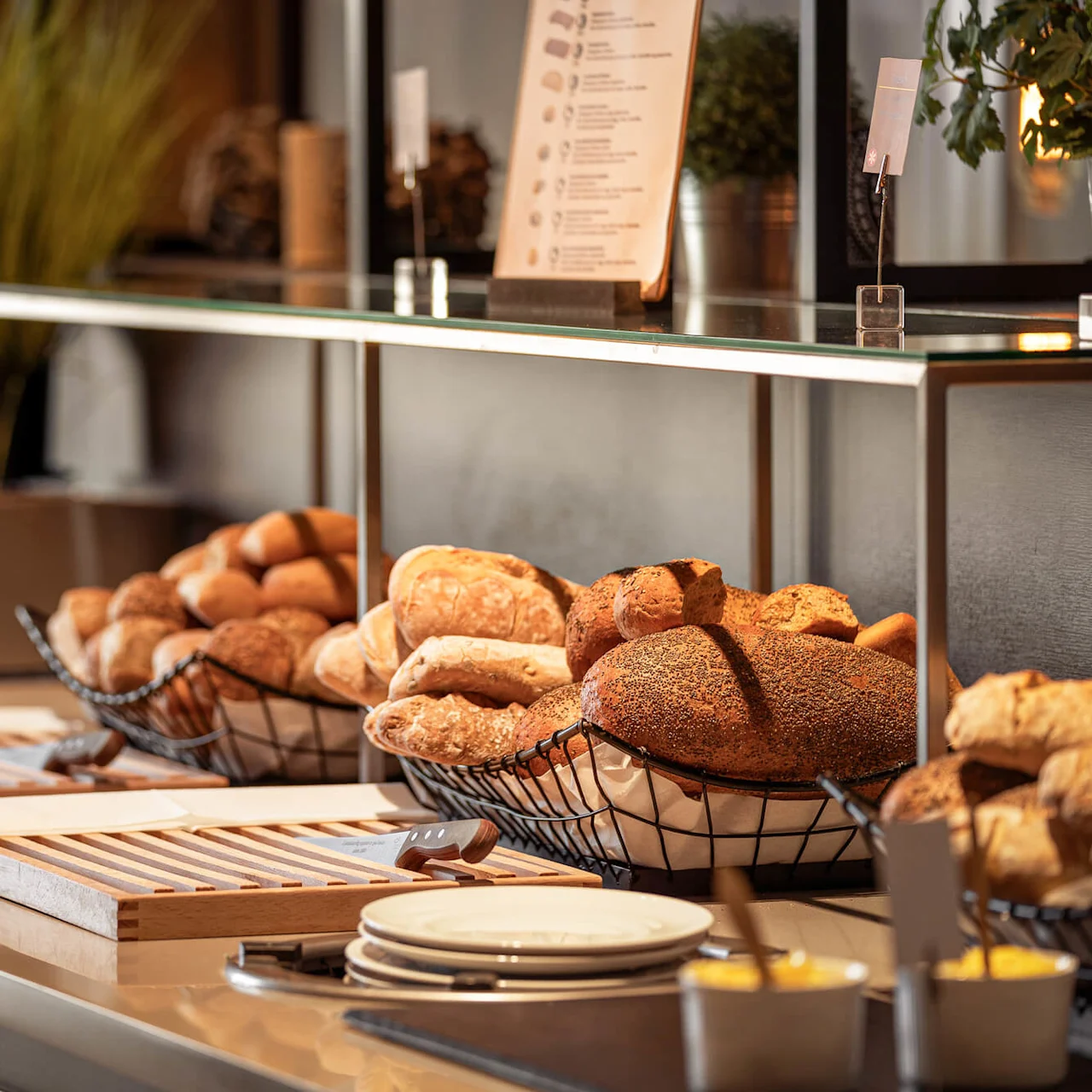 Baskets hold assorted breads on a countertop with cutting boards and butter dishes. Plants and a menu stand in the background.