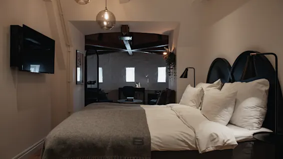 En snyggt bäddad säng med vita kuddar och en grå filt står i ett modernt sovrum med hängande lampor, en väggmonterad TV och en valvöppning till en mysig sittgrupp.