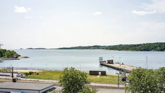 En naturskjønn kystutsikt med en lang brygge, rolig vann og grønne strender. En bygning ved navn Gamla Stationshuset.