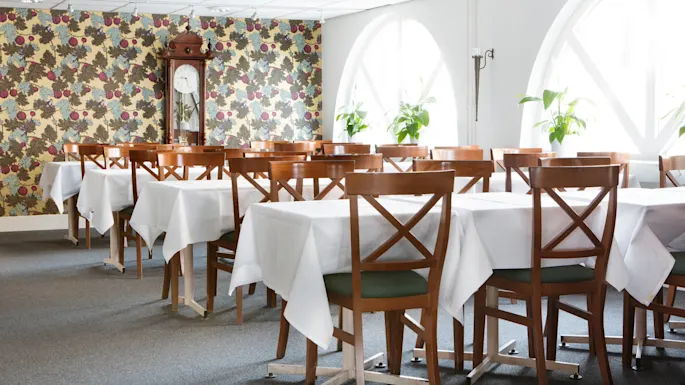 Trebord med hvite duker i en spisesal, sollys som trenger inn gjennom bueformede vinduer, mønstret tapet og et gulvur.