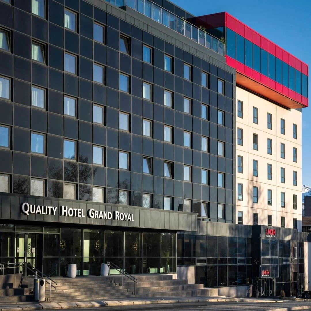 En moderne, fleretagers hotelbygning mærket "Quality Hotel Grand Royal" står fremtrædende. Ved siden af, komplementerer en rød struktur designet. Snedækkede bjerge er synlige i baggrunden.