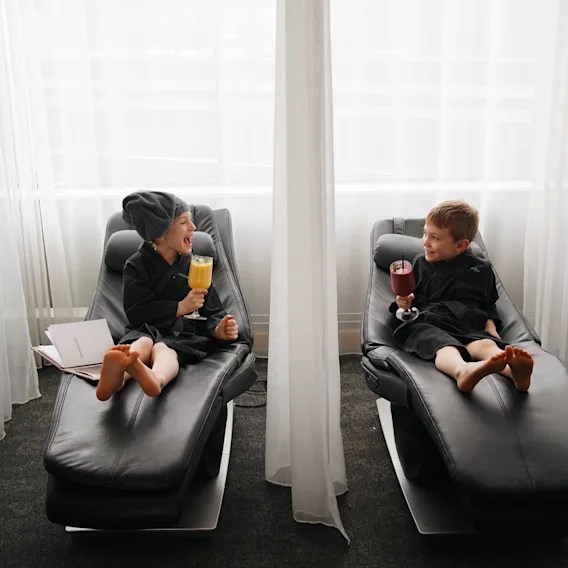 Two children in bathrobes relaxing on lounge chairs and enjoying colorful drinks at a spa