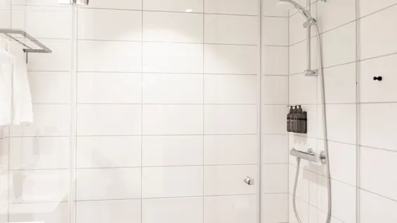 Valoisa ja puhdas moderni suihku valkoisilla kaakeliseinillä, lasiovella ja seinään kiinnitetyillä hygieniatuotteilla, käyttövalmiina.
