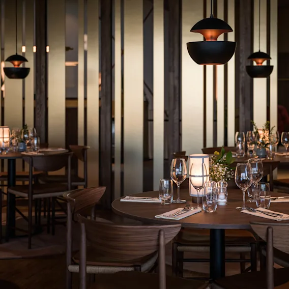 Bordene er elegant dekket med glass og servietter, opplyst av mykt stearinlys i en svakt opplyst, moderne restaurant. Treavskillere og hengende lamper forsterker den koselige stemningen.