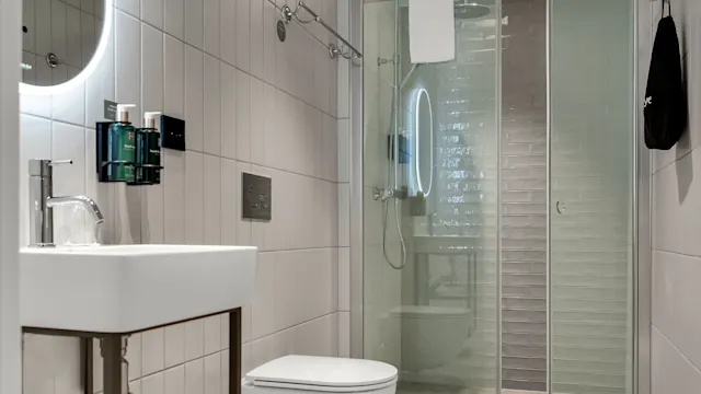 A modern bathroom with a white toilet, sink, mirror with backlight, and a transparent shower stall, with beige tiles.