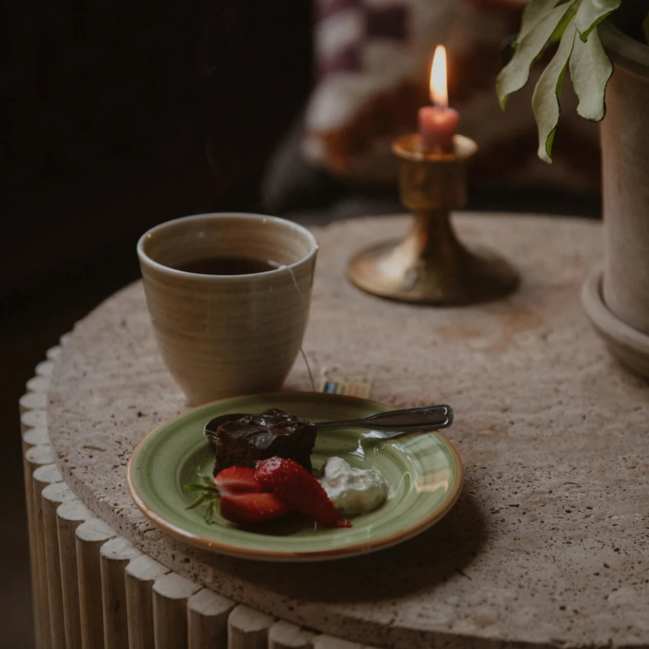 Cup of tea on a round table with a plate containing cake, strawberries, and cream. A lit candle and plant in the background create a cozy atmosphere. 