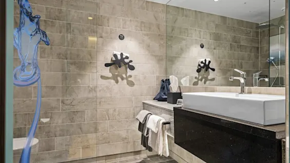 A modern bathroom features a rectangular white sink on a dark counter. Towels hang nearby, with mirrored walls reflecting decorative elements. Beige tiles cover the walls and floor.