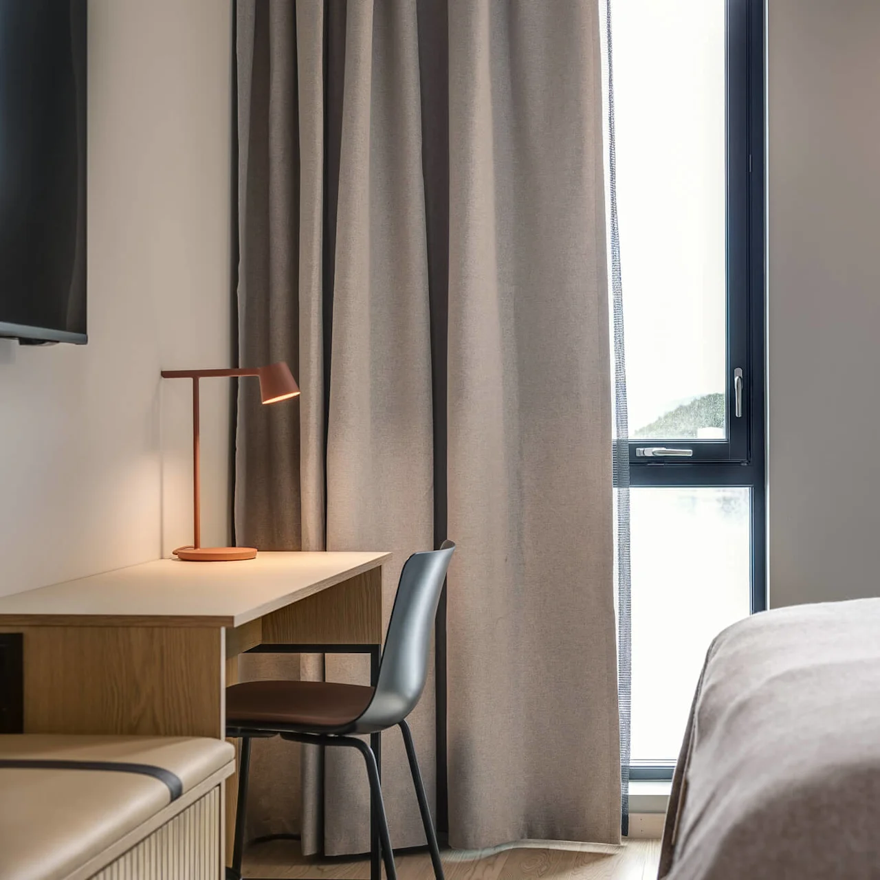 Et veloplyst hotelværelse med skrivebord, stol og lampe ved vinduet. Gardiner dækker delvist vinduet og afslører udsigt til et bjerg og vand udenfor.