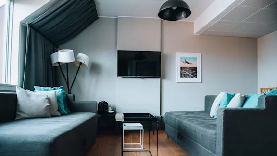 To grå sofaer med turkise puder er placeret over for hinanden i en moderne stue. Et sort TV er monteret på væggen over et lille glas sofabord.