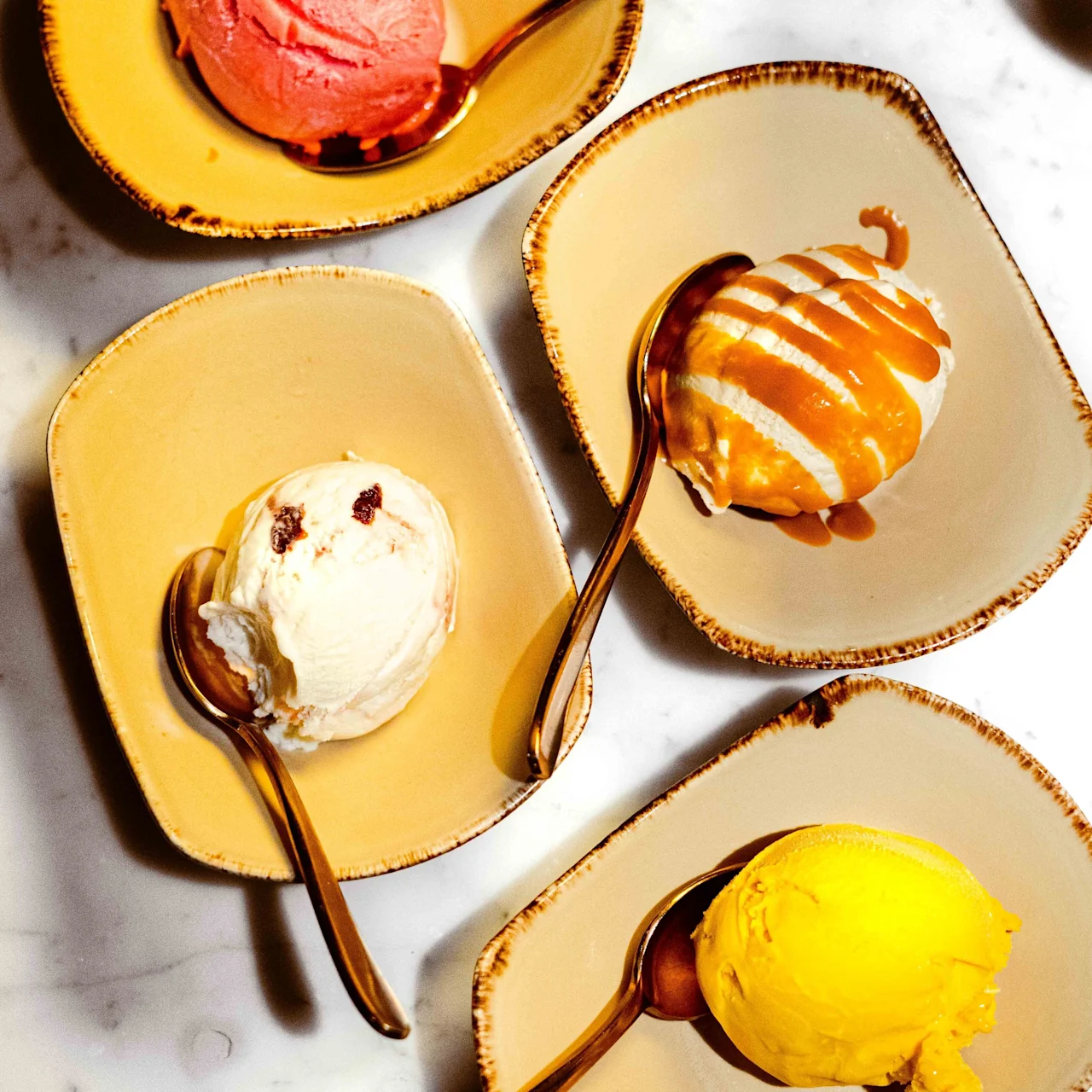 Four different ice cream scoops, each in a separate shallow bowl with a spoon, rest on a marble surface. Flavors appear to be strawberry, vanilla, caramel, and mango.
