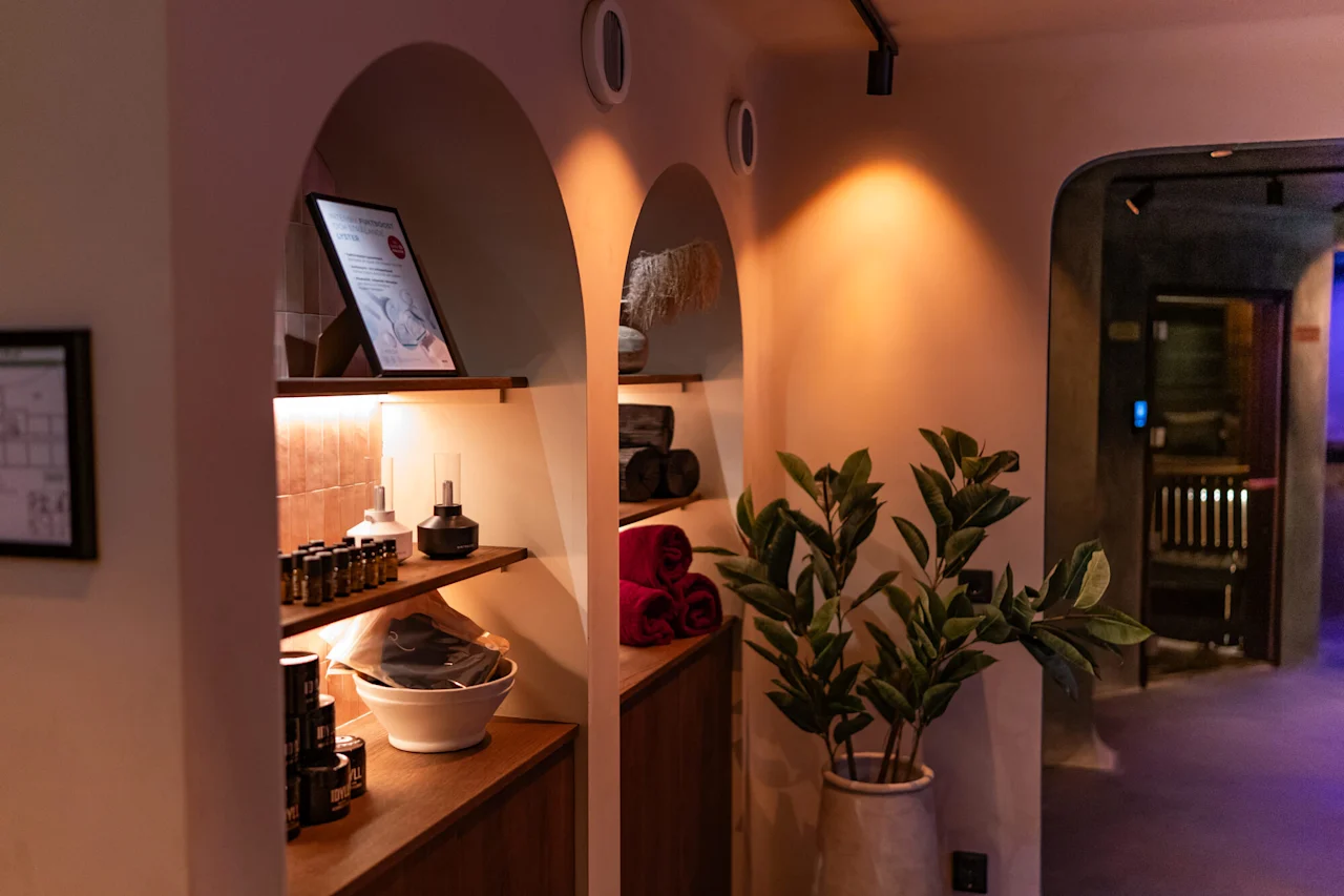 Viihtyisä kylpylä- tai hyvinvointialue, jossa on kaarevia syvennyksiä, lämmin valaistus, hyllyjä tuotteineen ja pyyhkeineen sekä tietoa tabletilla.