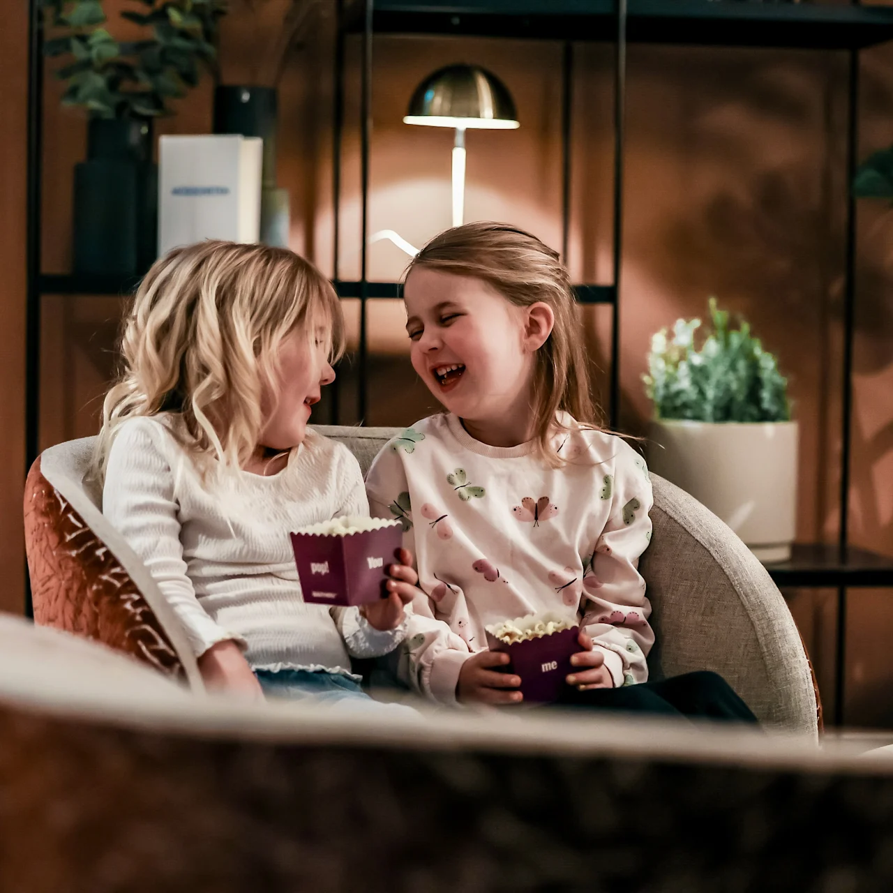 Two children sit close on a couch, laughing and holding small popcorn boxes. The room is warmly lit, with plants and a lamp in the background.