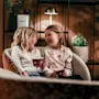 Two children sit close on a couch, laughing and holding small popcorn boxes. The room is warmly lit, with plants and a lamp in the background.