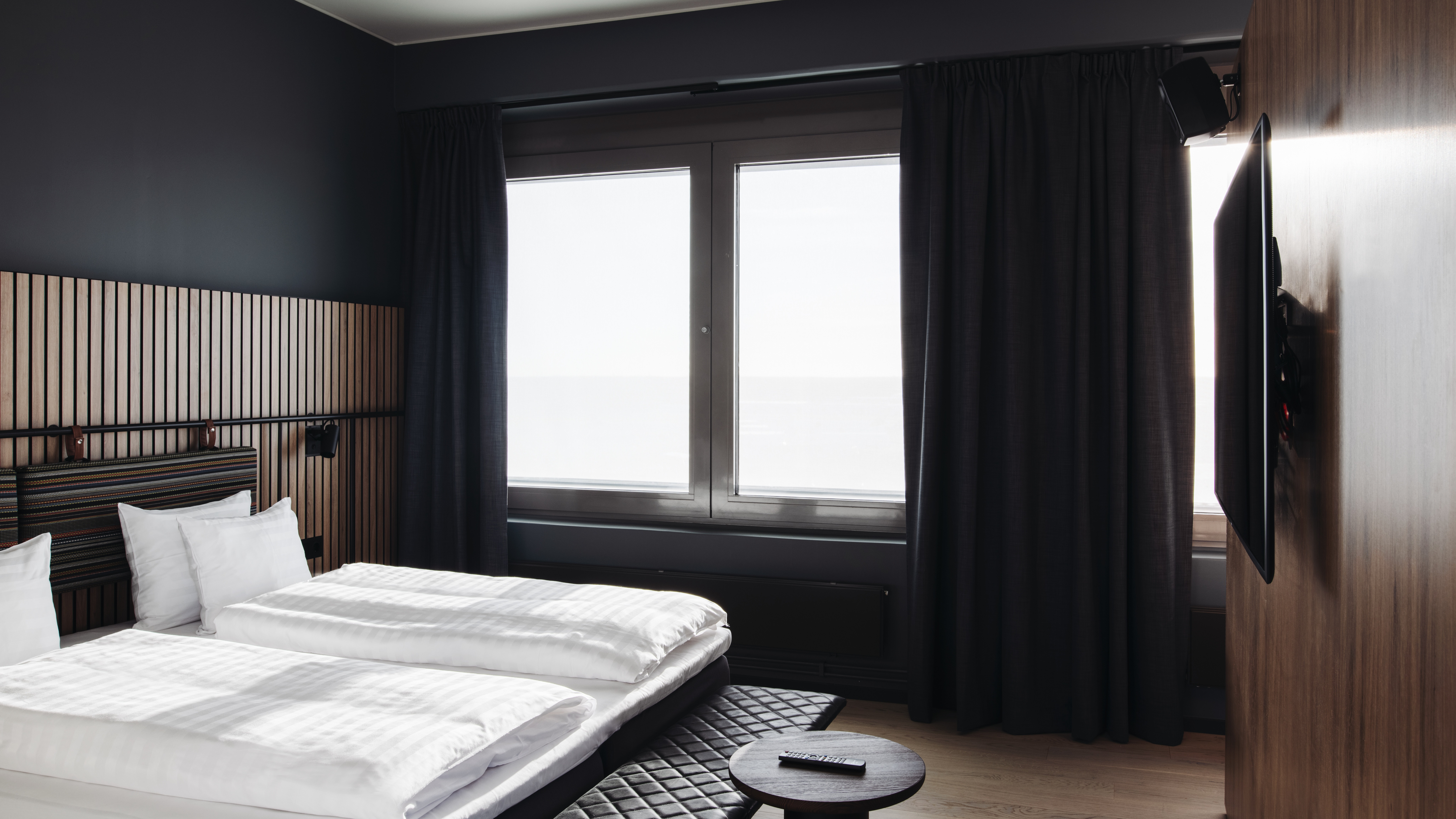 Et moderne hotelværelse med to senge, mørke gardiner og et stort vindue, der slipper lyst ind.