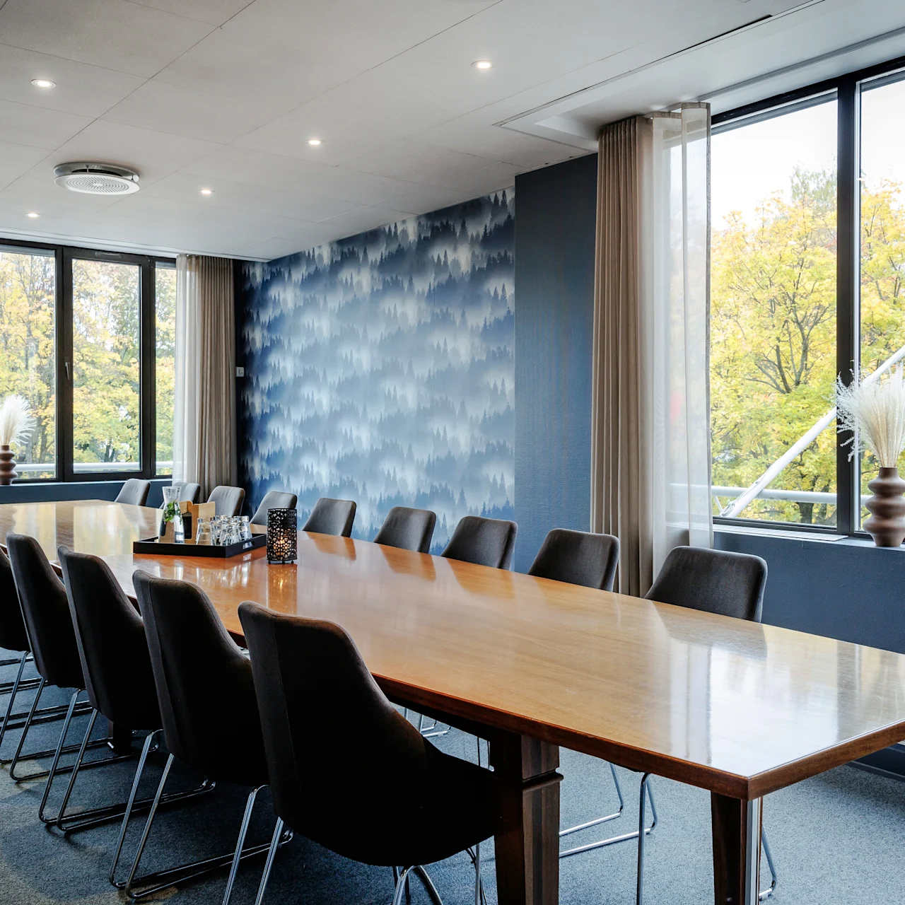 A long wooden table surrounded by black chairs sits in a conference room. Large windows reveal trees outside, and decorative vases with white flowers accent the space.