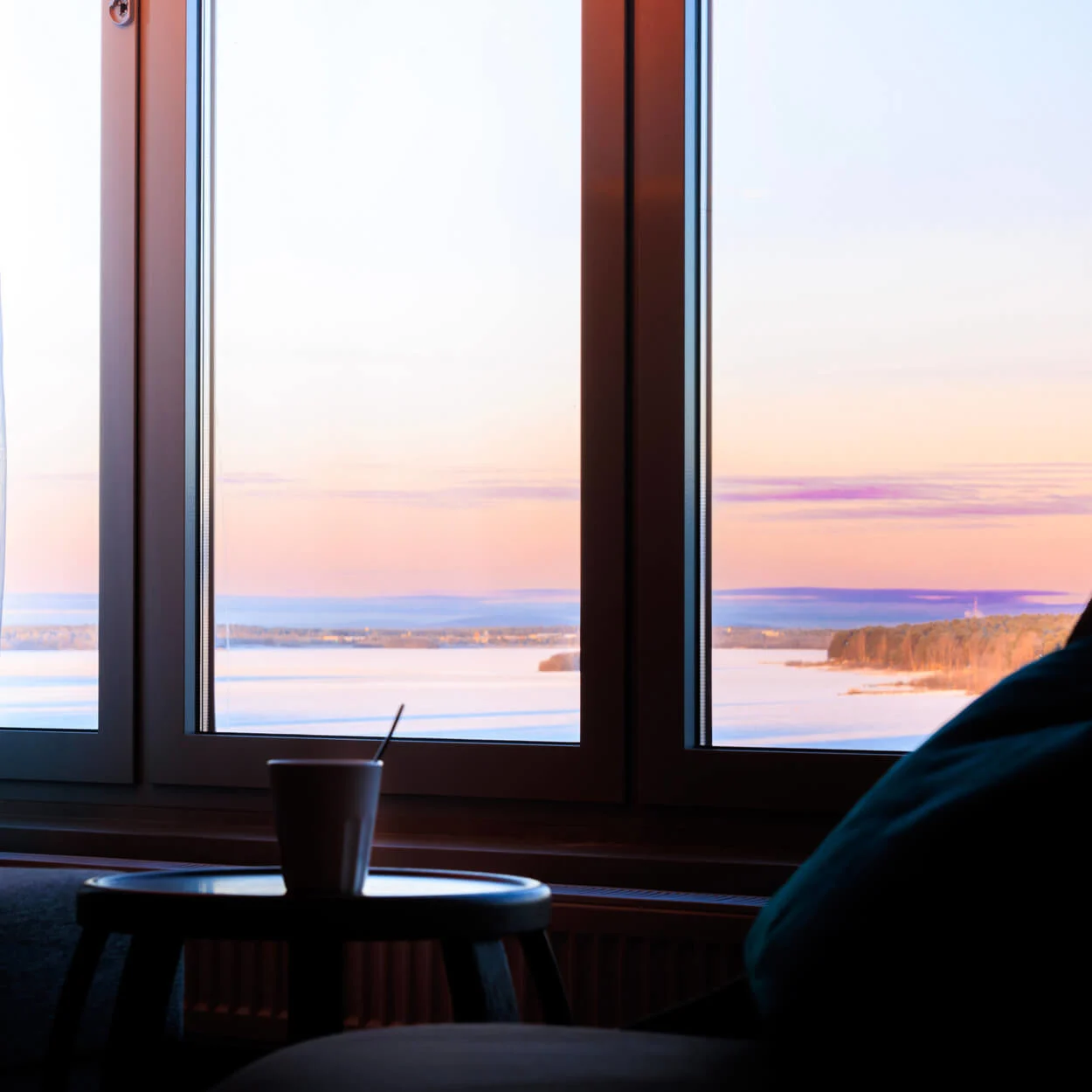 Ett mysigt rum med en kopp på ett litet bord framför ett stort fönster, där genomskinliga gardiner ramar in en naturskön vy av en färgstark solnedgång över vatten.