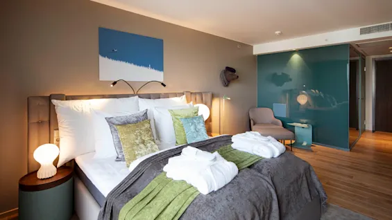 A neatly made bed with pillows and robes sits in a modern hotel room. Soft lighting, art on the wall, and a seating area create a comfortable, stylish atmosphere.