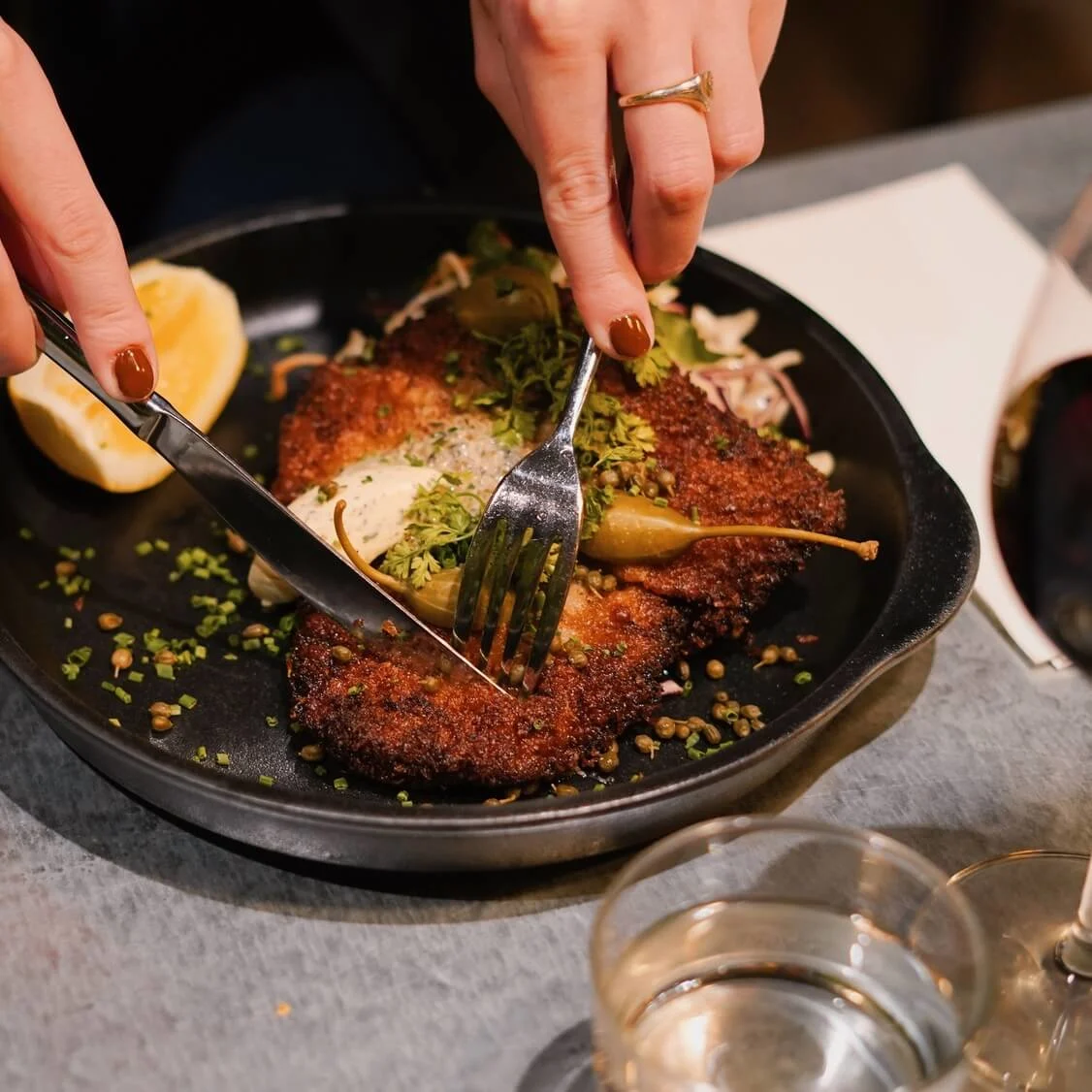 A breaded cutlet is cut with a fork and knife. Garnished with herbs and accompanied by lemon, it’s served on a black plate. Nearby, there's a glass of red wine and water.