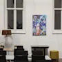 Två höga lampor ramar in en färgglad målning på en vit vägg mellan två fönster. Nedanför står bord och stolar i en stilren, modern inomhusmiljö.