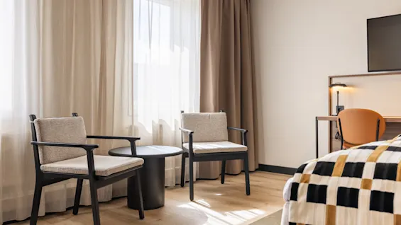 Valoisa hotellihuone, jossa kaksi nojatuolia ja pieni pöytä ikkunan ääressä, sänky kuviollisella peitolla ja työpöytäalue.