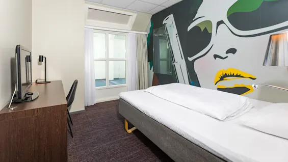 A small hotel room features a bed with white linens, a desk with a TV and lamp, and a large, colorful mural of a face with sunglasses on the wall.