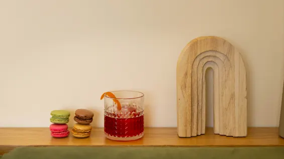 Glas innehållande röd dryck med apelsinskal, stående på en trähylla bredvid färgglada macarons och en träbåge, med minimalistisk konst och torkad växt i bakgrunden.