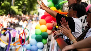 Folk jublar och vinkar från en paradvagn, med färgglada ballonger längs kanten. Den omgivande miljön är fylld av liv och rörelse, vilket antyder en festlig händelse.