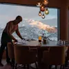 En person ordner et stort spisebord med glass og bestikk, inne i et koselig rom med hengende lys, mot en bakgrunn av snødekte fjell synlige gjennom store vinduer.