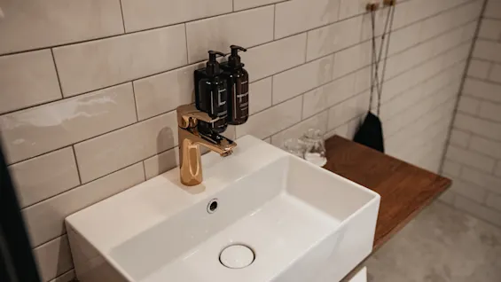 En moderne badeværelsesvask med guldkran, hvide fliser og vægmonterede sæbe- og kropsvaskdispensere.