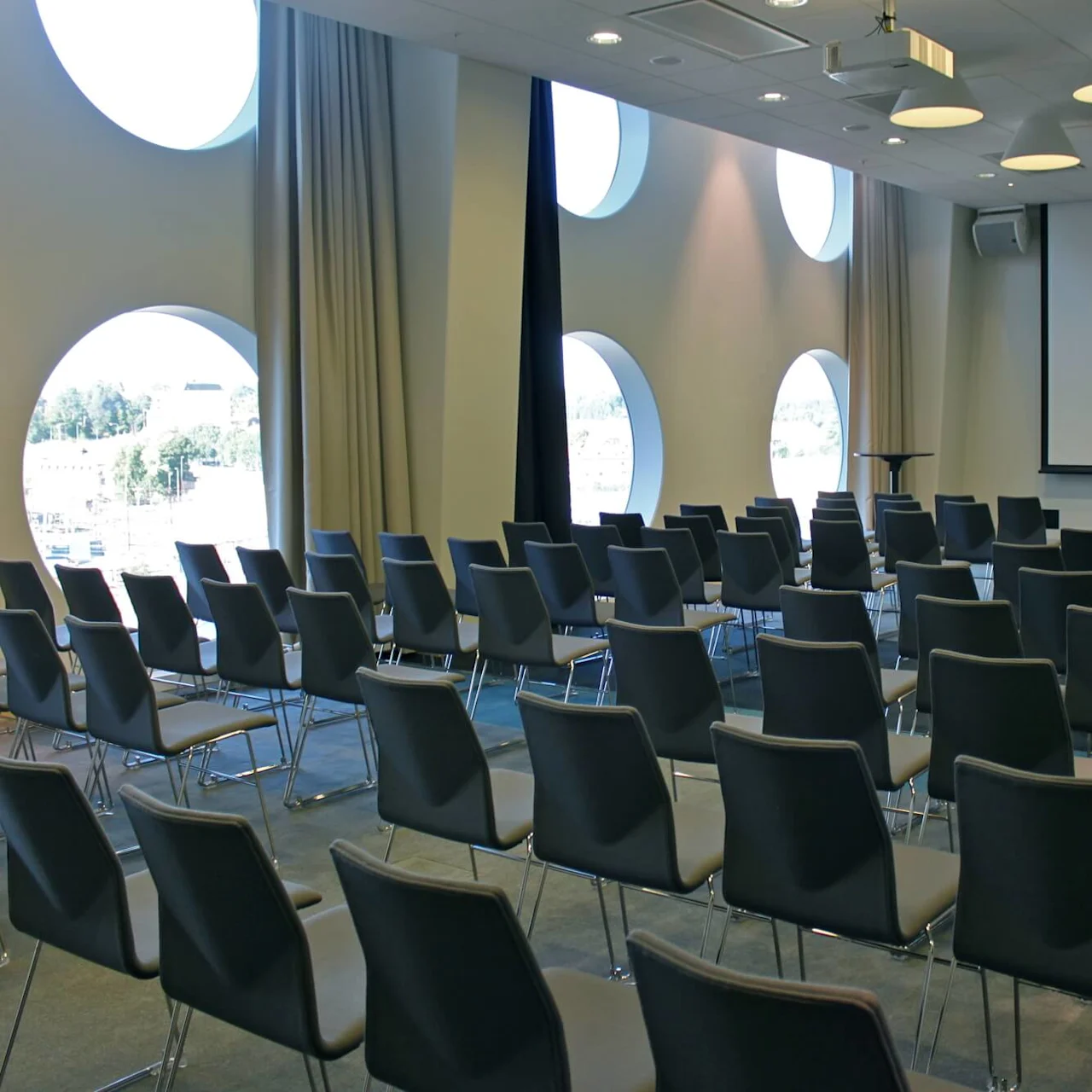 Rækker af tomme sorte stole vender mod en stor tom skærm i et moderne konferencelokale med runde vinduer og beige gardiner.