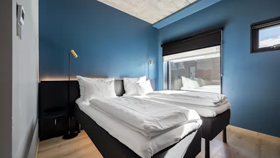 Et moderne hotellrom med to komfortable senger med hvite laken, blå vegger og et stort vindu for naturlig lys.