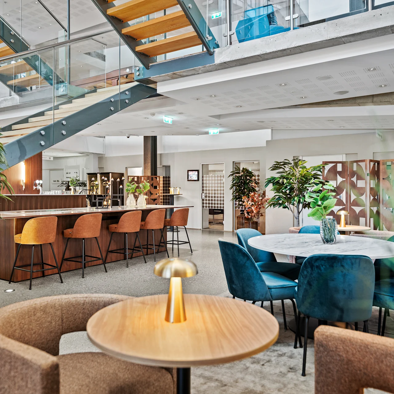 En modern lounge med runda bord och stoppade stolar. Miljön har en trappa och en träbar med pallar, omgiven av växter och mjuk belysning.