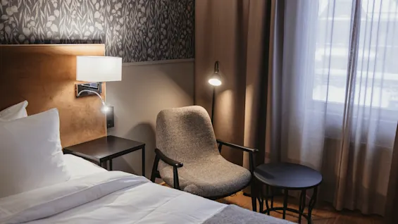 Ett mysigt hotellrum med säng, fåtölj och varm belysning, med mönstrad tapet och ett fönster med skira gardiner.