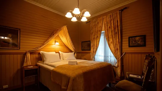 A large bed with white pillows and a yellow coverlet stands under a soft light in a warmly lit, vintage-style bedroom with wooden walls and patterned curtains.