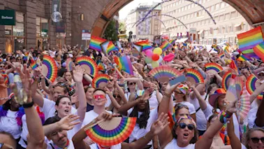 En livlig folkemengde feirer i en parade, vifter entusiastisk med regnbueflagg og vifter under en bybue.