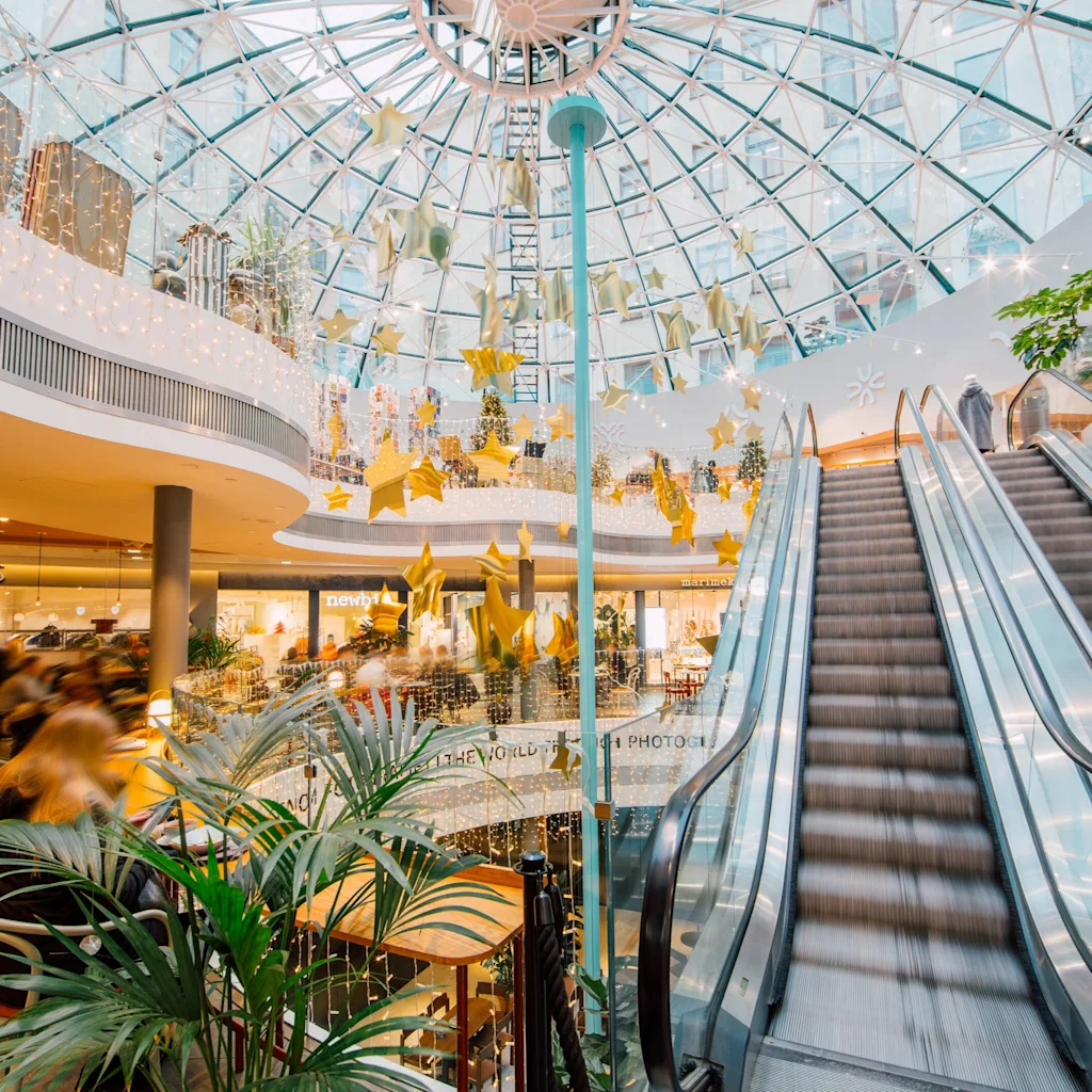 Inne i Kämp Galleria i Helsingfors, ett livligt köpcentrum med glaskupol, rulltrappor och festliga stjärndekorationer.