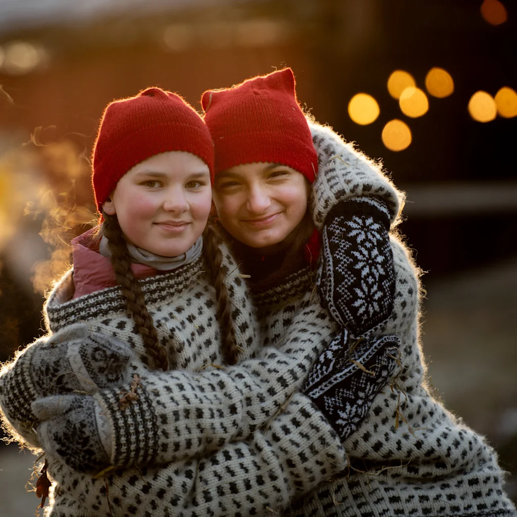 Two people in red hats and warm sweaters embracing outdoors with warm lights in the background.