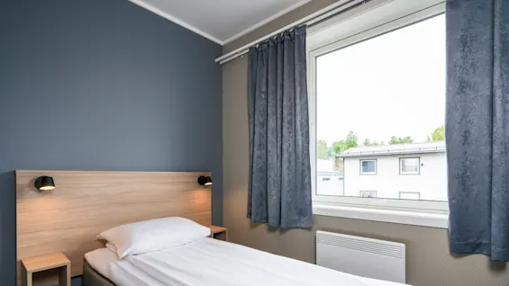 A neatly made single bed rests against a blue wall, illuminated by small wall lamps, in a bright room with a large window and gray curtains.