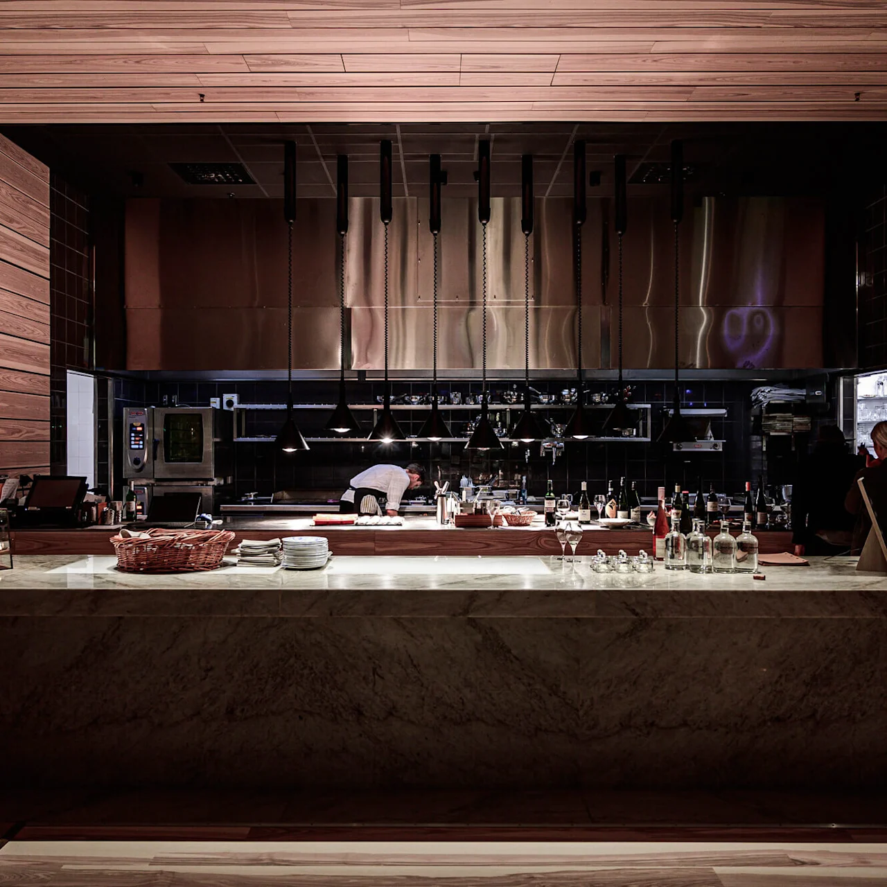A chef works in a modern, open kitchen with wooden accents and sleek equipment. People dine nearby, while bottles and tableware sit on the counter, creating a cozy atmosphere.