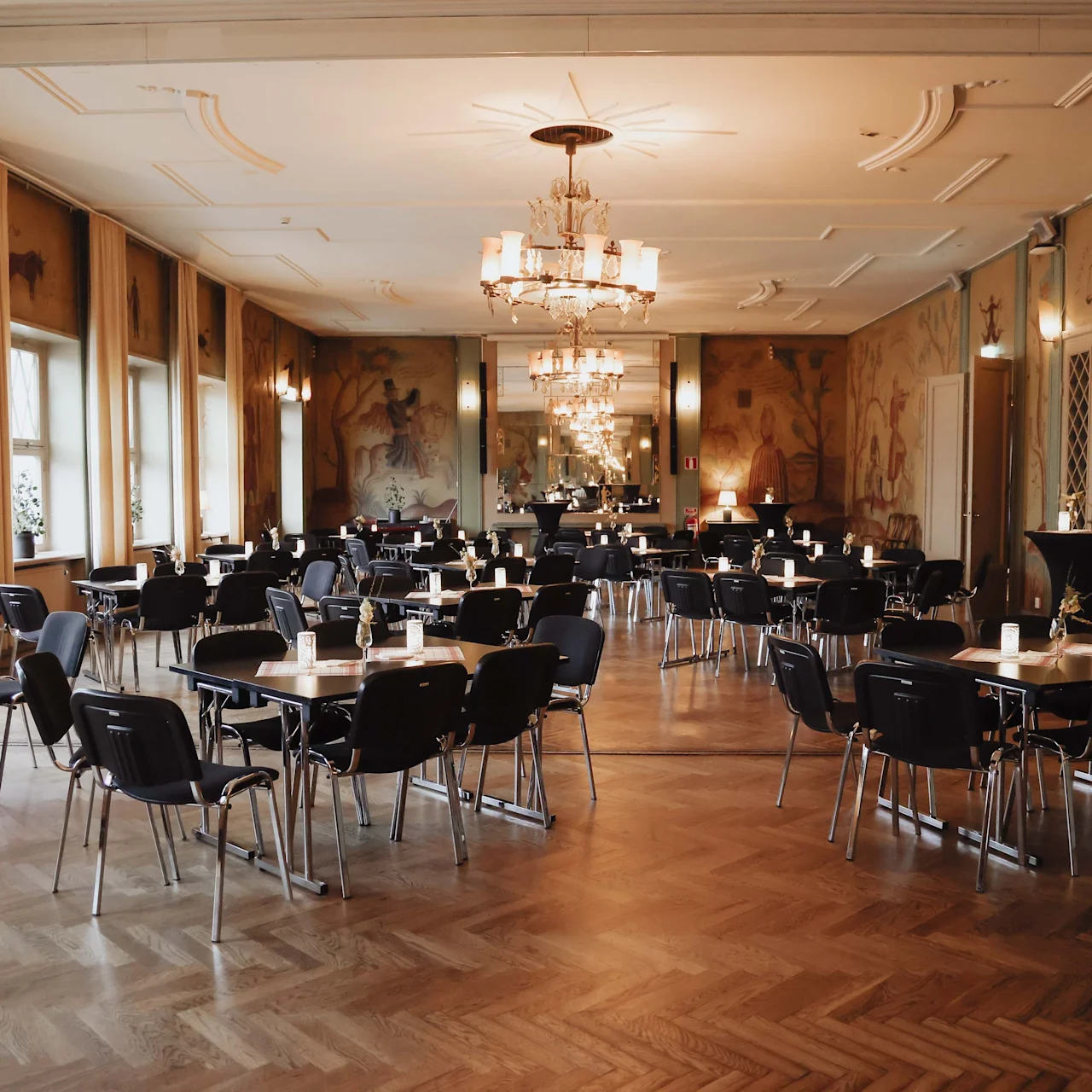 Tables and chairs are arranged neatly in an elegant, spacious room with chandeliers and patterned wall art. Soft lighting creates a warm, inviting atmosphere, complemented by large windows.