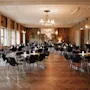 Tables and chairs are arranged neatly in an elegant, spacious room with chandeliers and patterned wall art. Soft lighting creates a warm, inviting atmosphere, complemented by large windows.