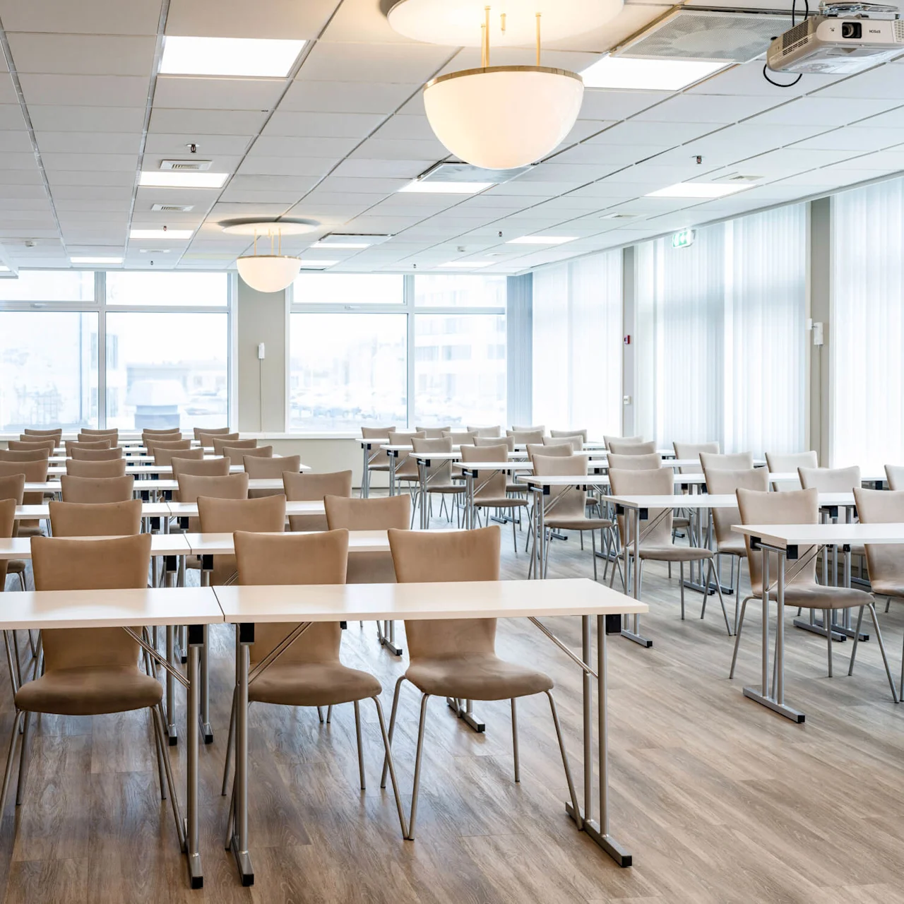 Chairs and tables arranged in rows fill a brightly lit conference room with large windows, wooden flooring, and ceiling lights. A projector hangs from the ceiling.