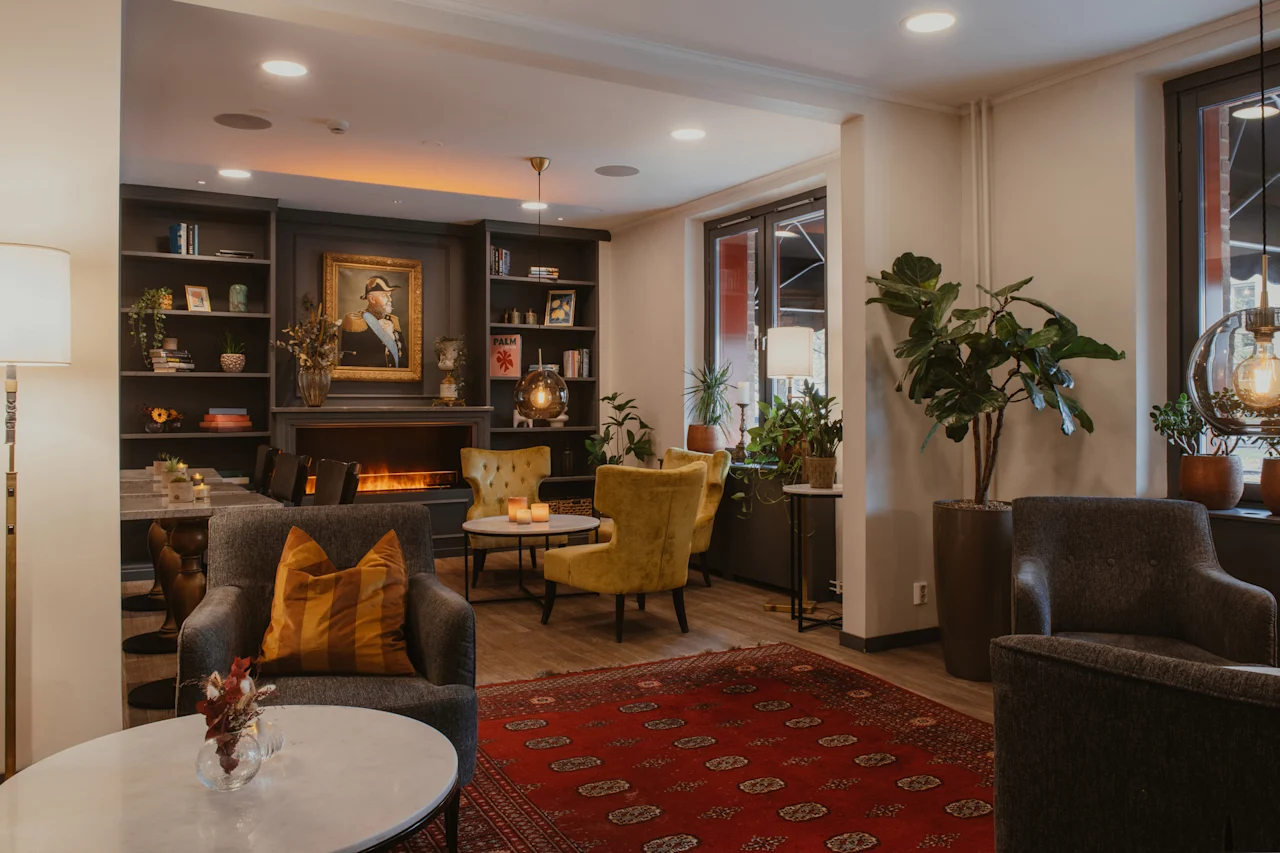 A cozy hotel lounge with comfortable seating, a fireplace, and bookshelves, featuring PALM. Home Hotel Kung Oscar.