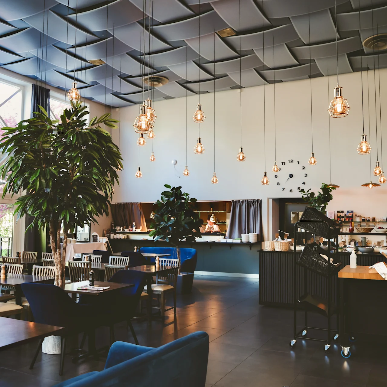 Elegant café interior; trees and hanging lights create ambiance over tables and chairs. Counter in the background displays pastries and beverages under a stylish, patterned ceiling.