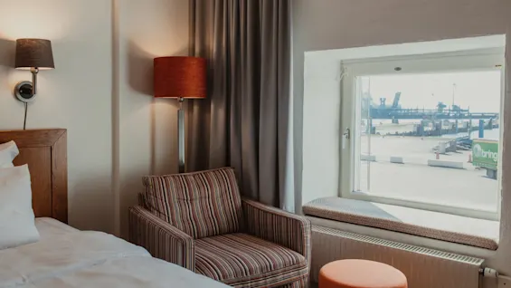 Viihtyisä hotellihuone, jossa on sänky, raidallinen nojatuoli ja ikkuna, josta avautuu näkymä vilkkaaseen satamaan.